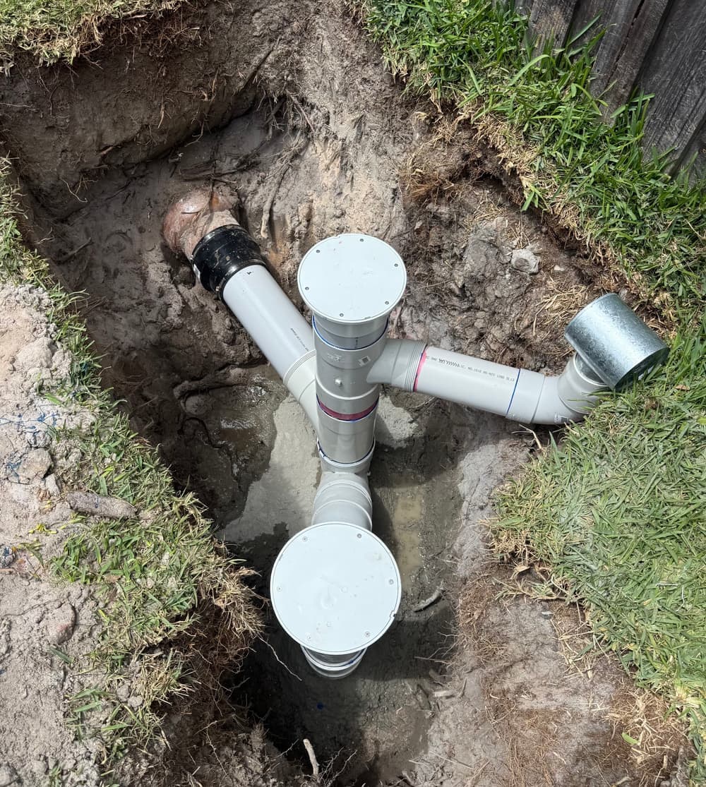 Excavated backyard trench showing a new sewer junction with inspection openings connected to existing earthenware pipework