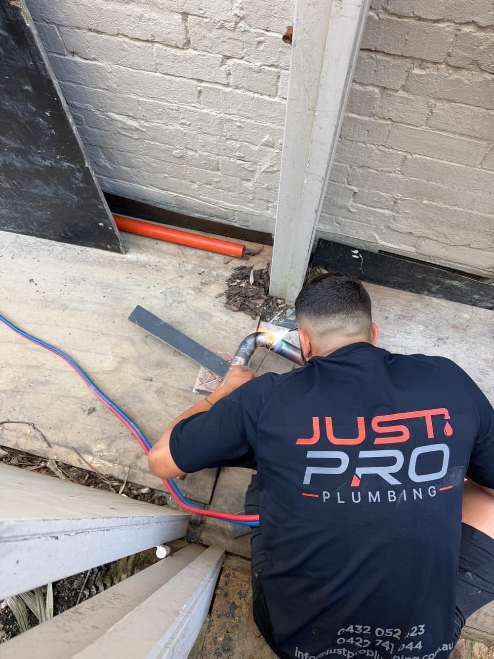 Just Pro Plumbing technician in branded workwear brazing a copper pipe joint beneath a deck, beside a brick wall